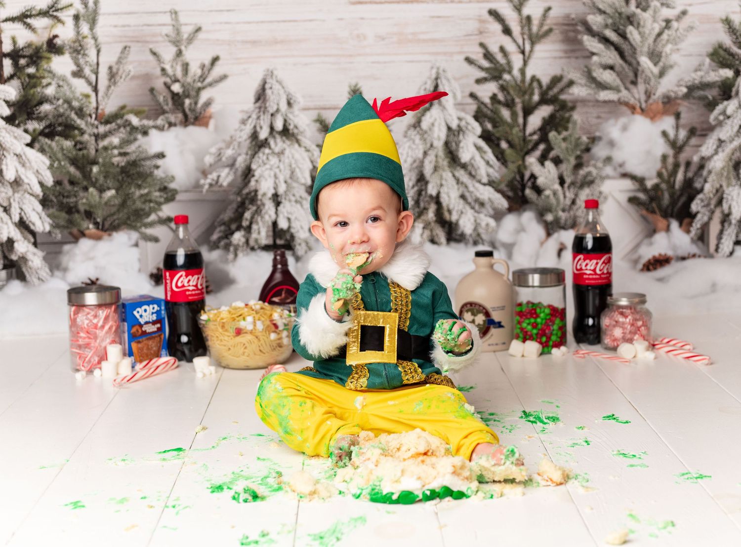 Baby in elf costume sits among Coca-Cola bottles and snowy trees during festive Christmas cake smash photo session. cake smash photographer