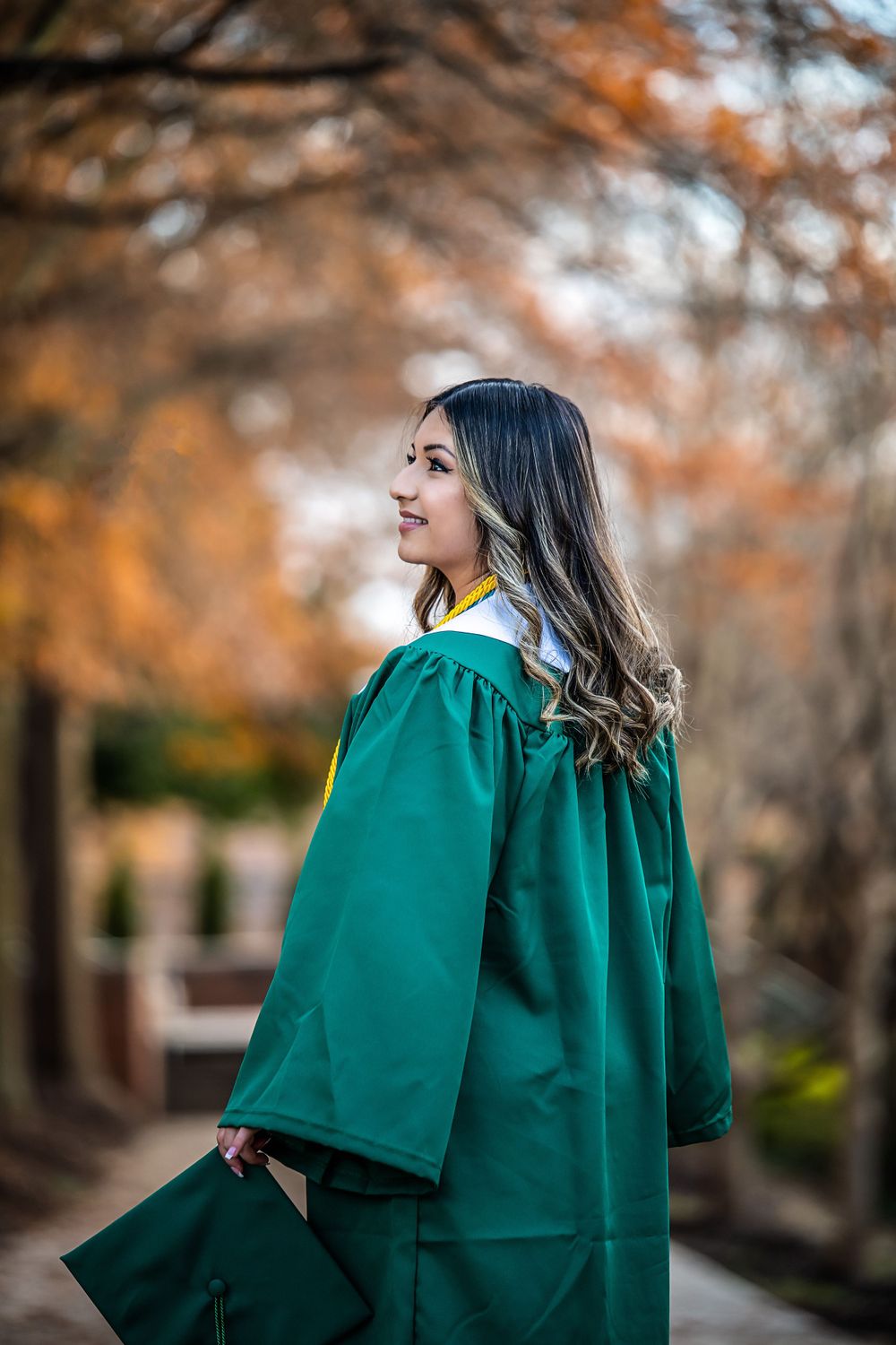 Graduate in emerald green academic regalia poses against autumn foliage background.