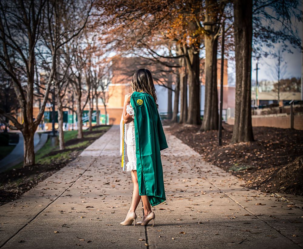 Graduate walking along tree-lined path at sunset in emerald green graduation gown.