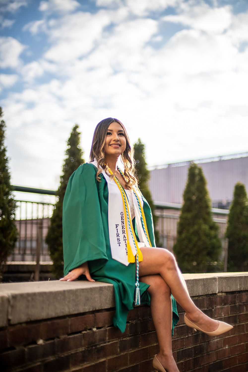 Graduate in green cap and gown sitting casually on brick wall under blue sky.