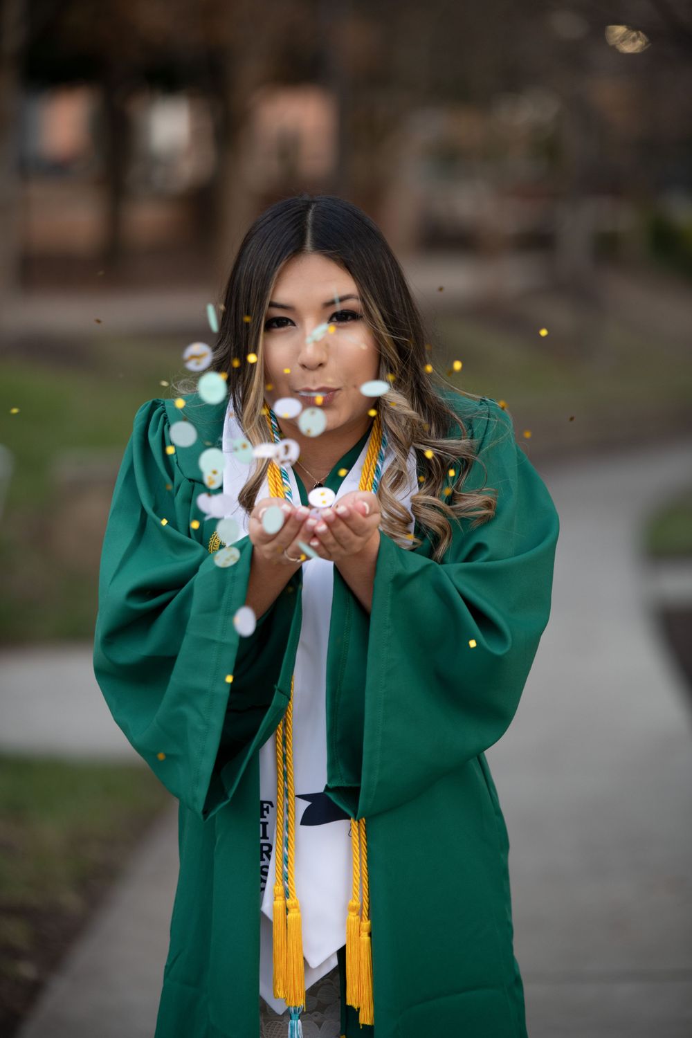 Graduate in green gown blowing glitter from hands in magical graduation portrait.
