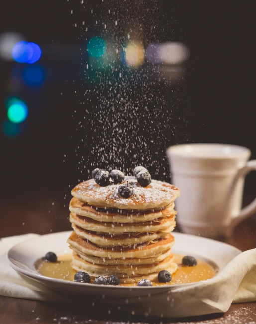 Stack of fluffy pancakes with blueberries and powdered sugar being dusted from above.