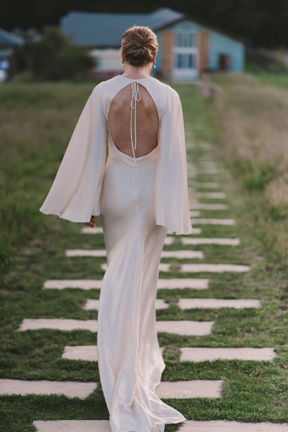 White wedding dress with flowing cape sleeves shown from behind on stone pathway.