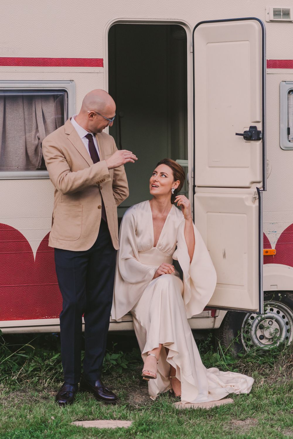 Couple sharing a moment outside vintage camper with bride in white dress and groom in beige suit.