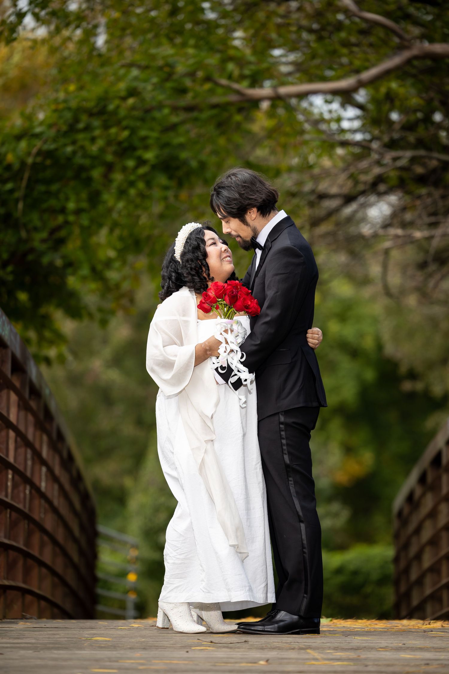 A romantic wedding couple shares an intimate moment on a wooden bridge with red roses.