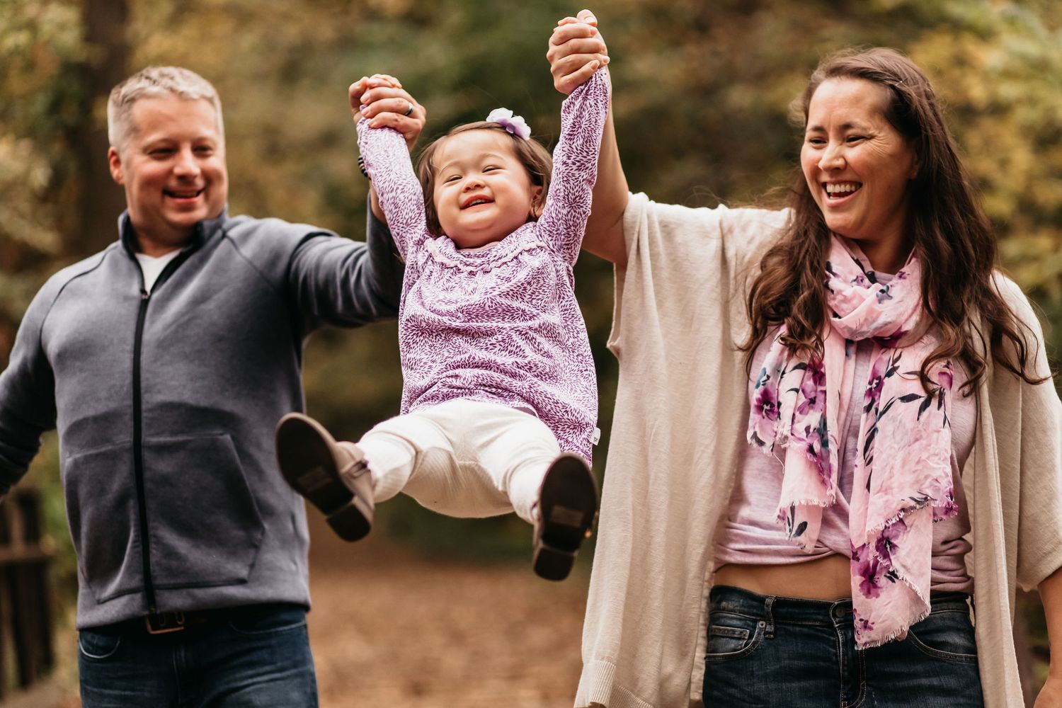A mom and dad hold their young daughter in between them as they swing her toward the camera. They're all laughing.
