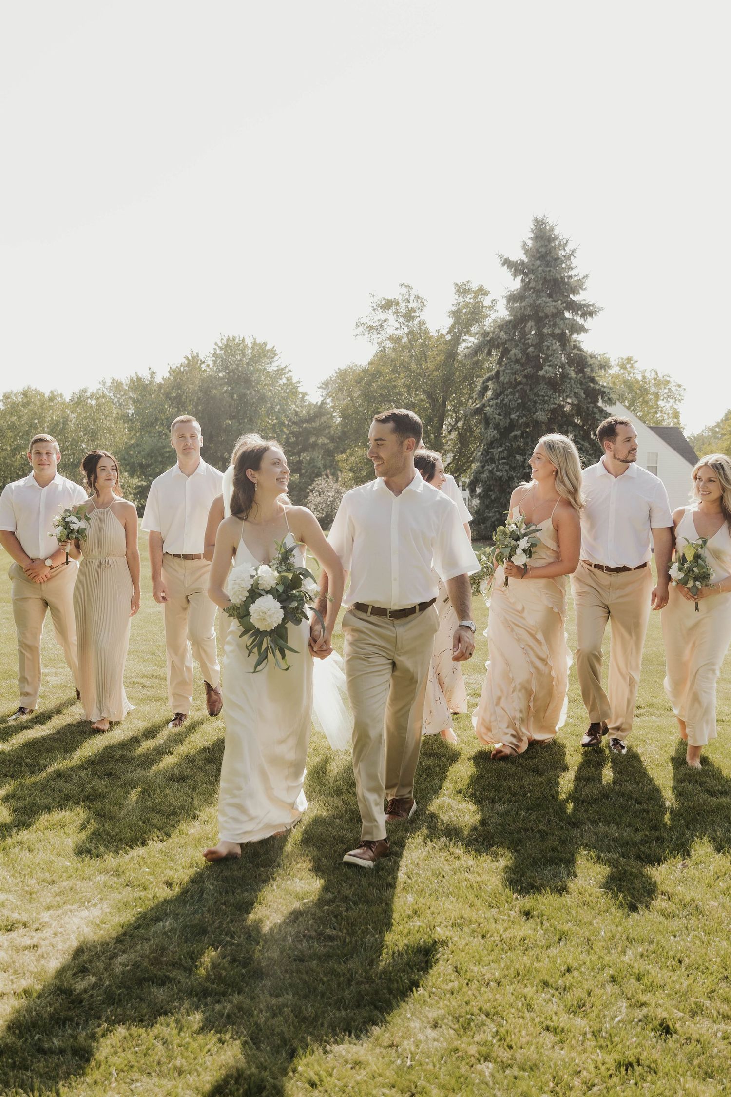 Wedding party walking together in white and beige attire through a sunny meadow.
