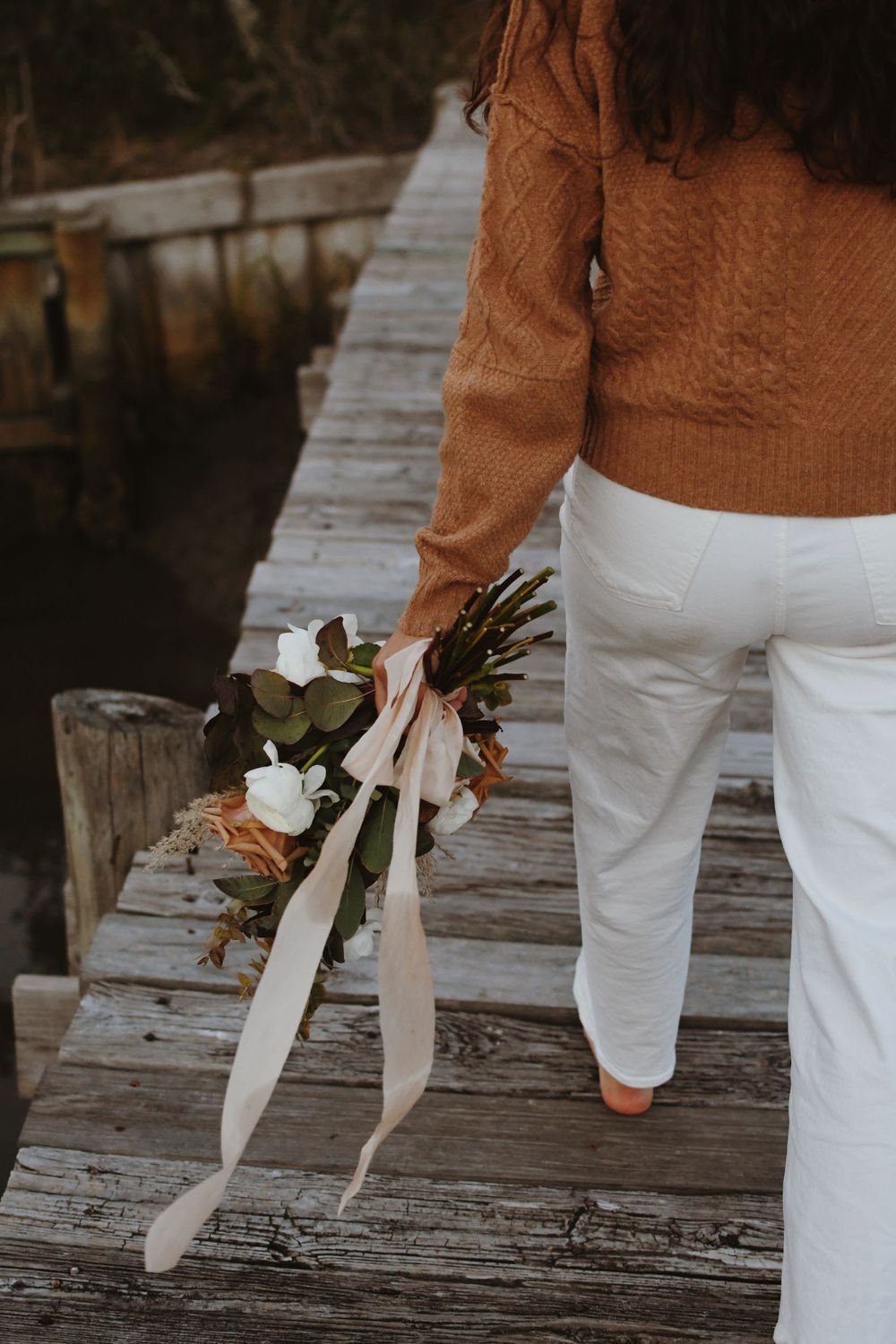 Person in white pants and brown sweater walking on wooden dock holding floral bouquet.