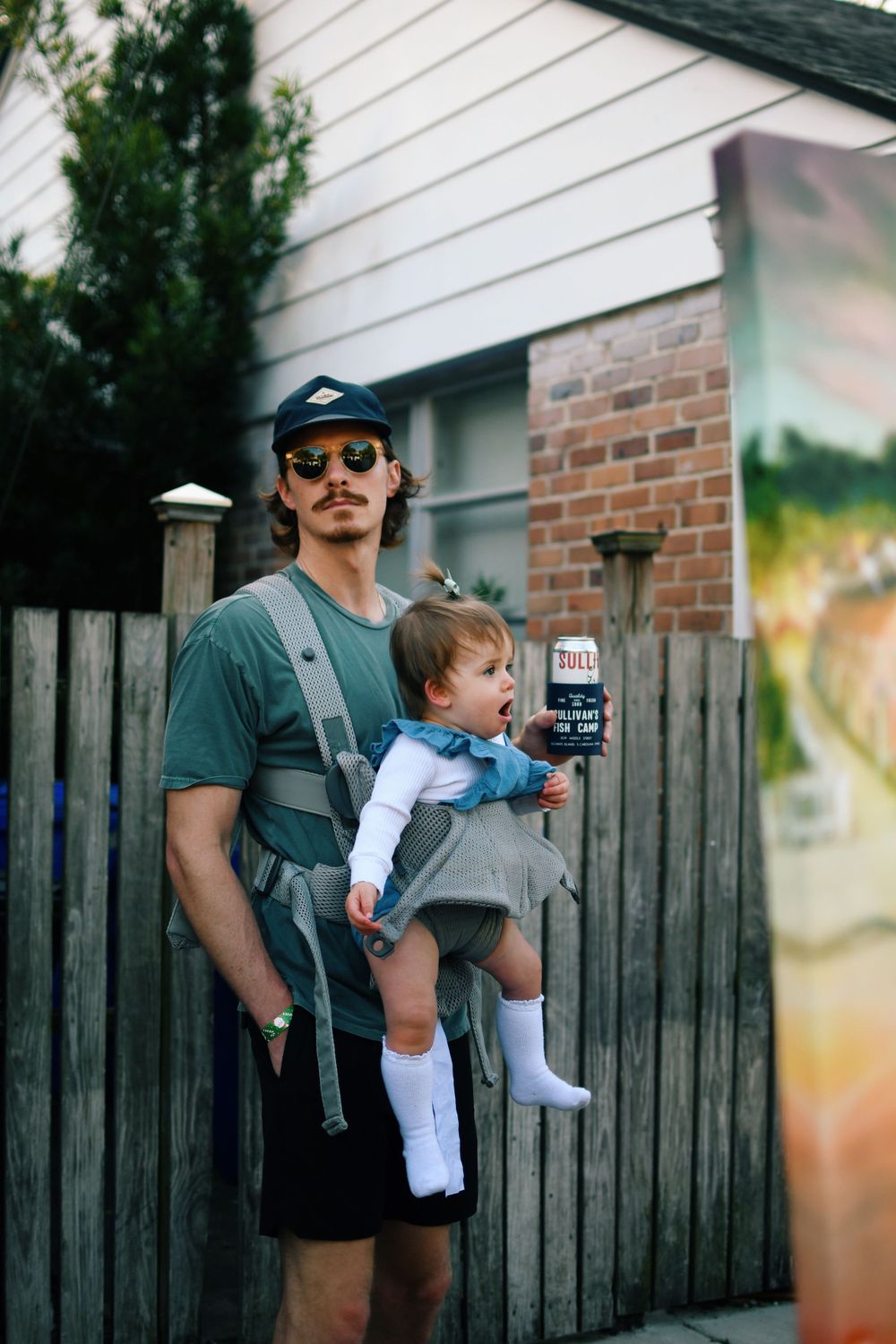 A person wearing sunglasses carries a small child in a baby carrier against a wooden fence backdrop.