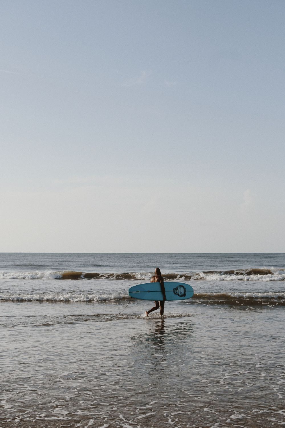 A lone surfer carrying a blue surfboard walks through ocean waves at sunset.