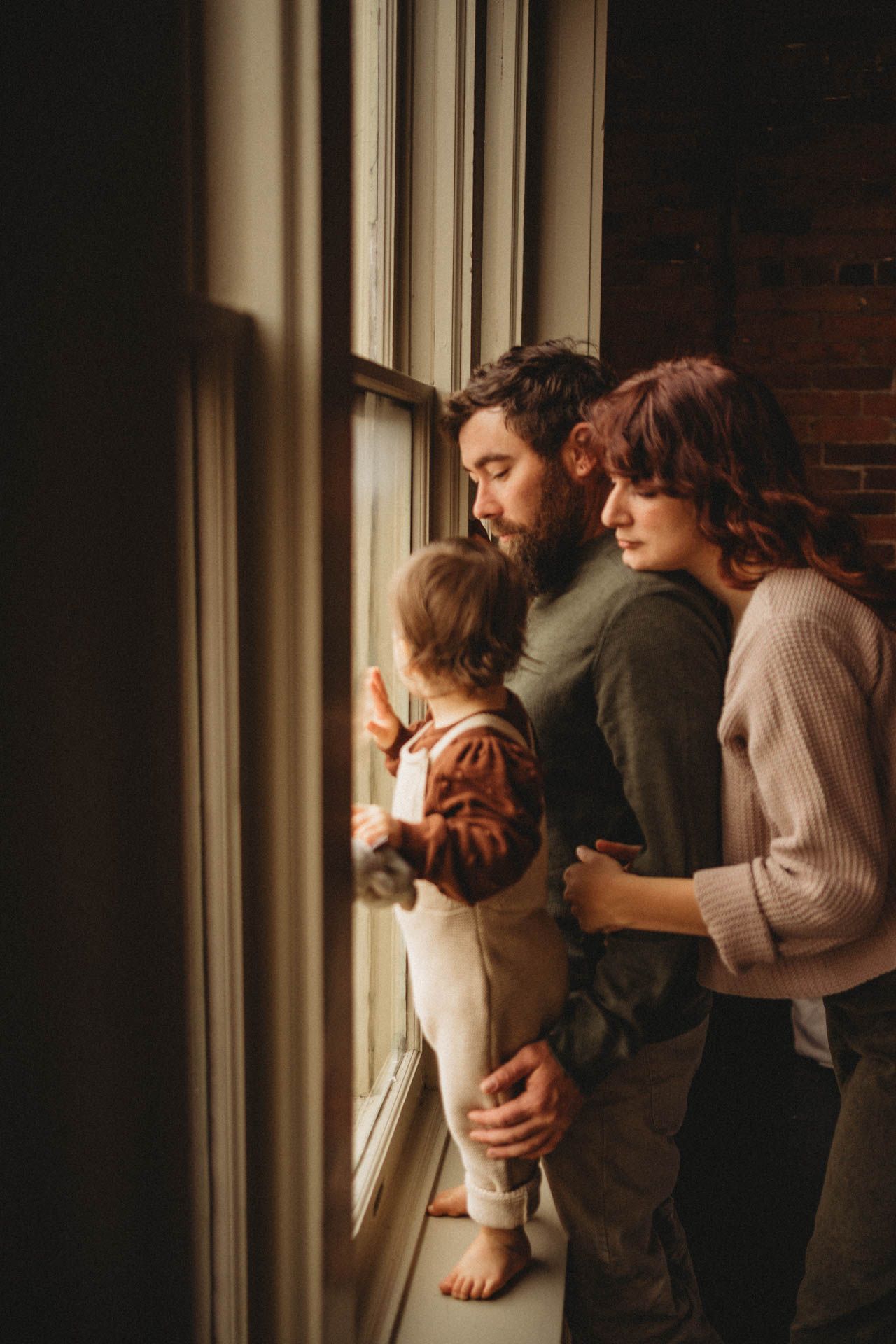 A tender family moment by a bright window in a rustic room with exposed brick walls.