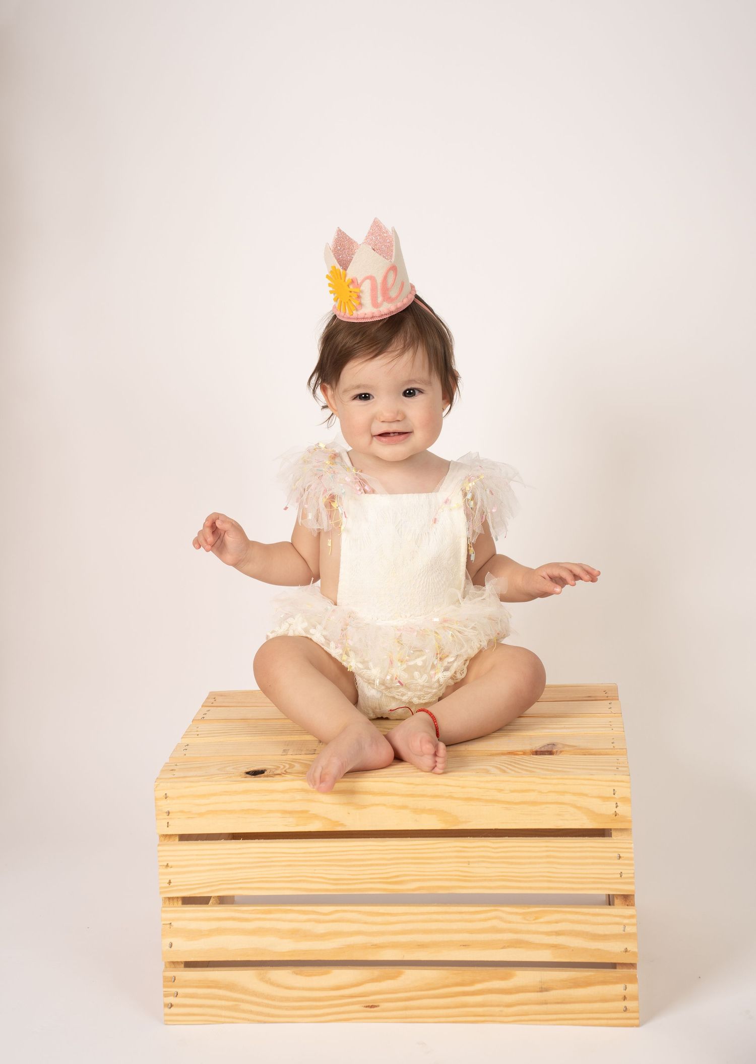 Baby in white outfit sitting on wooden crate during professional photo shoot.