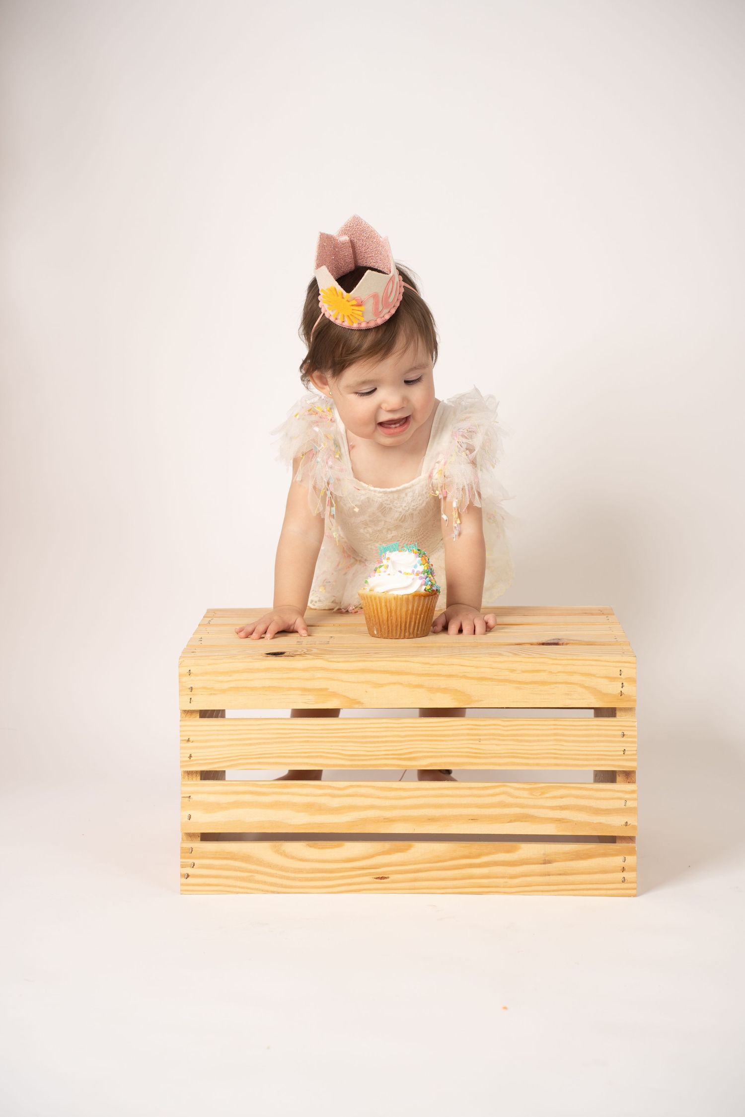 Baby in lace dress posing on wooden crate during photo session.
