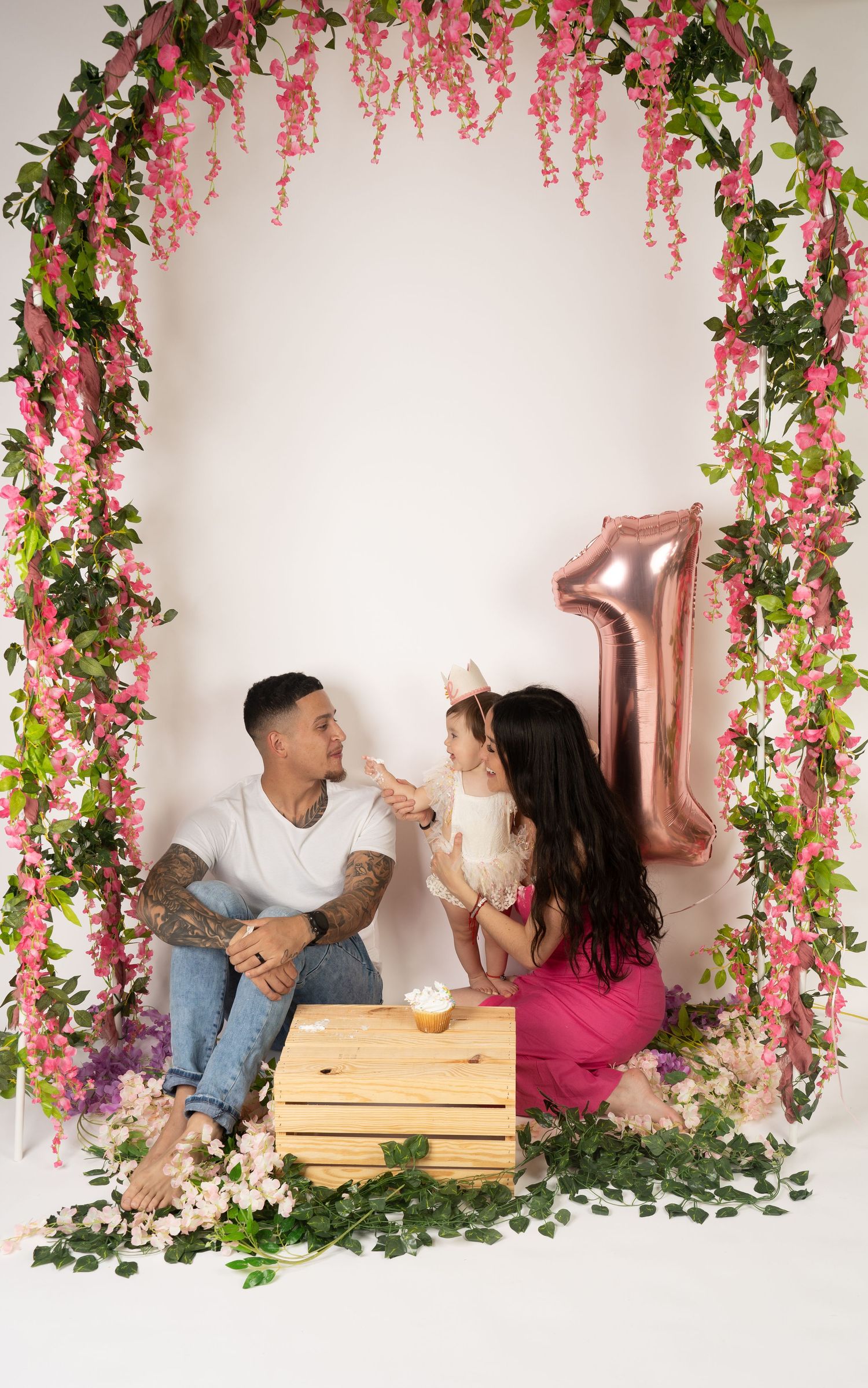 A romantic photo shoot setup with pink floral decorations and greenery hanging against a white backdrop.