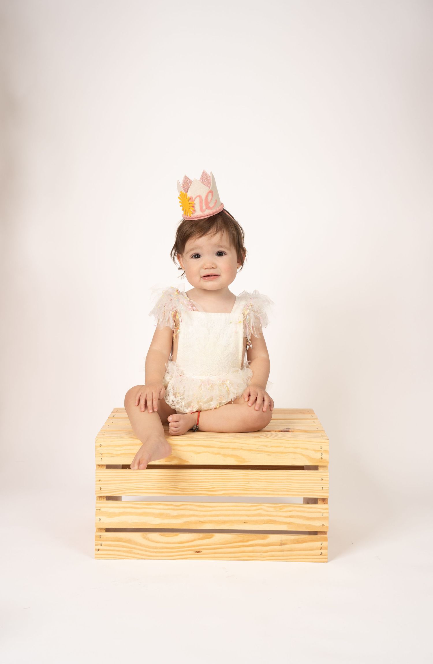 Baby wearing crown sitting on wooden crate against white backdrop.