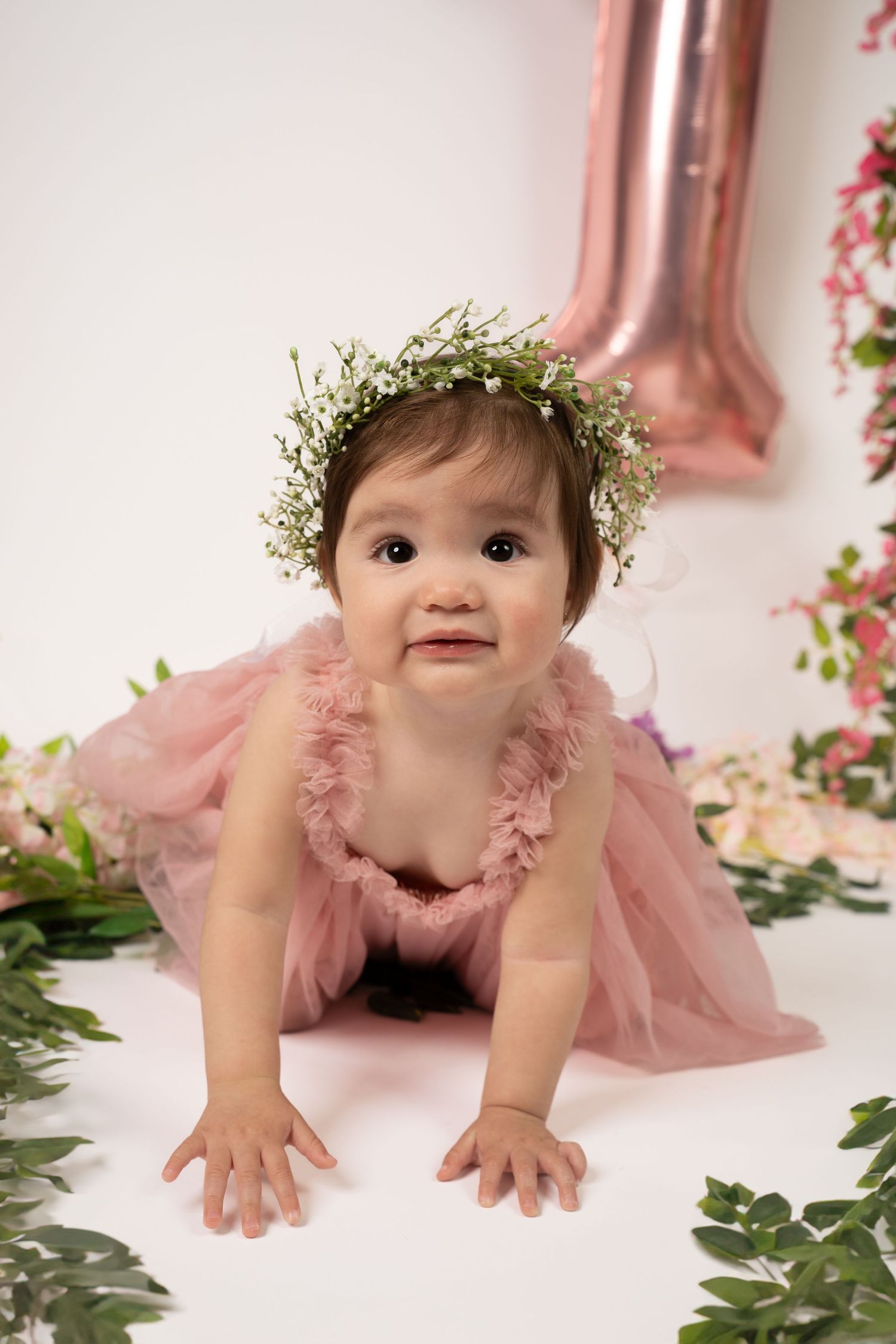 Baby wearing a pink tulle dress and floral crown crawls on white floor with pink decorations.
