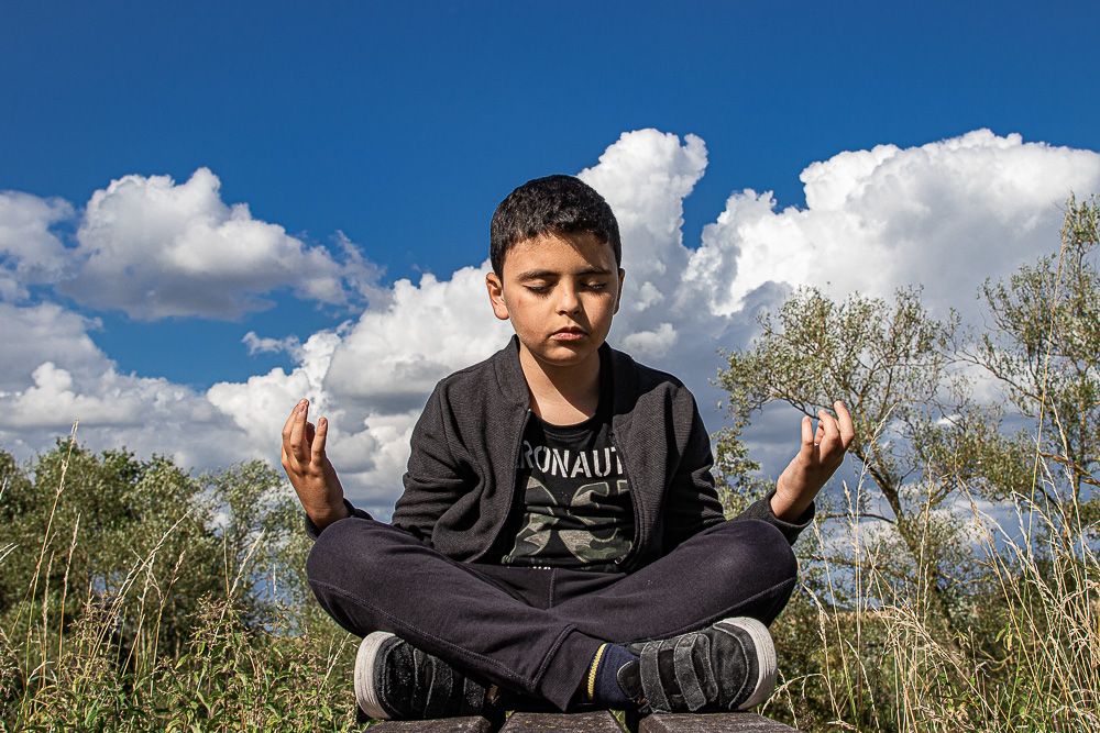 young boy sitting in meditation position on a bench in the green field with a blue sky with beautiful white clouds