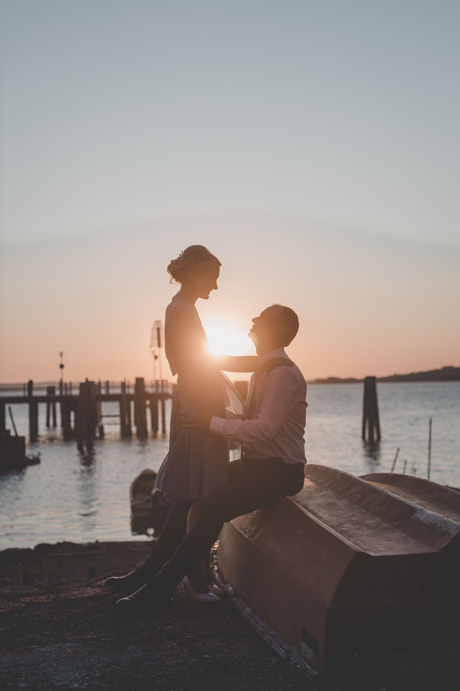 Couple sitting on beach dock at sunset creating romantic silhouettes against golden sky.