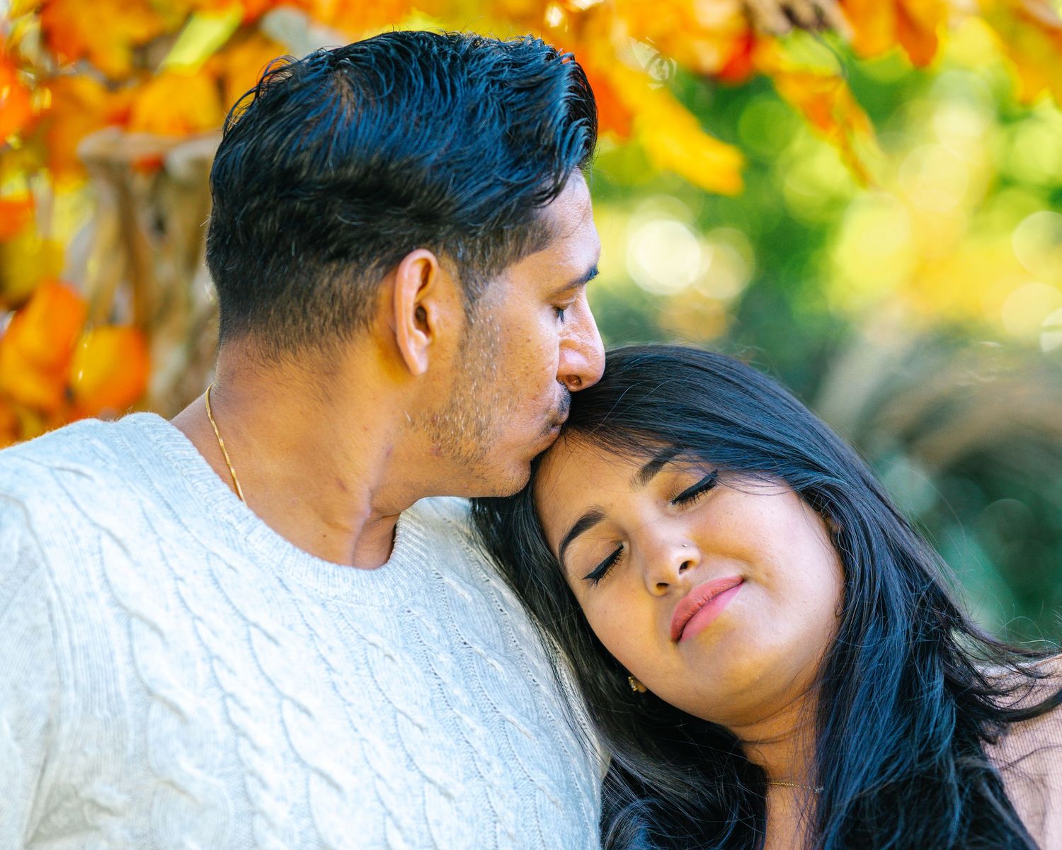 A couple shares an intimate moment outdoors with autumn foliage in the background.