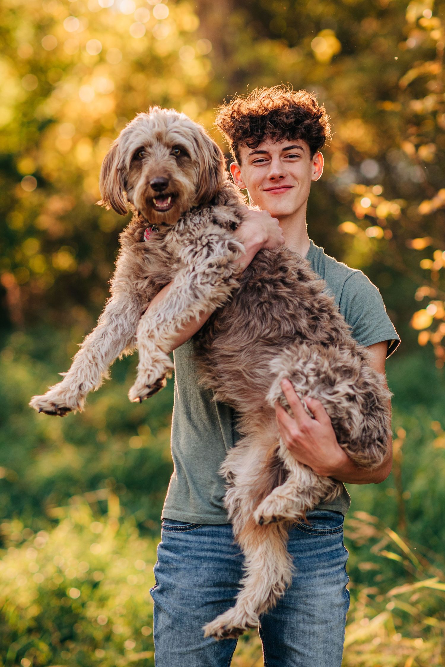 A young person holds a large fluffy goldendoodle dog outdoors during golden hour.