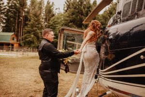 Wedding party standing together outdoors with forest scenery and rustic wooden structures.