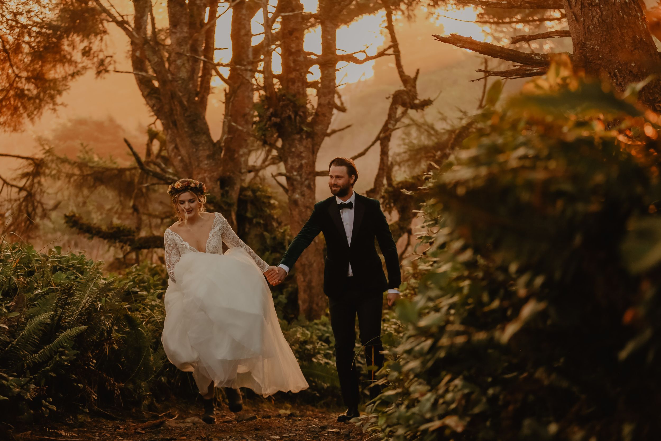 Couple walking through misty forest path in wedding attire during golden hour.