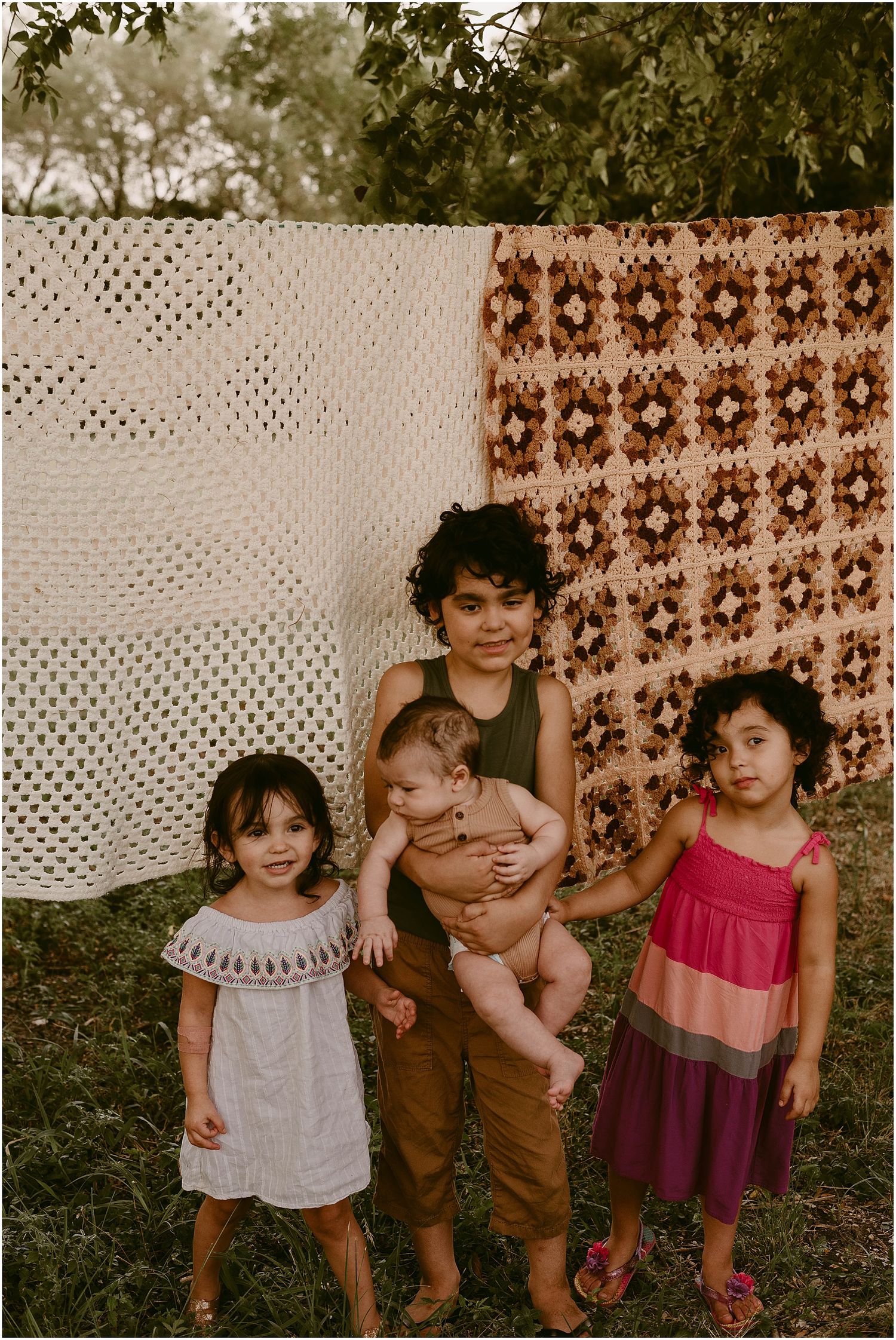 Three small children pose together against a backdrop of white crochet and brown patterned fabrics outdoors.
