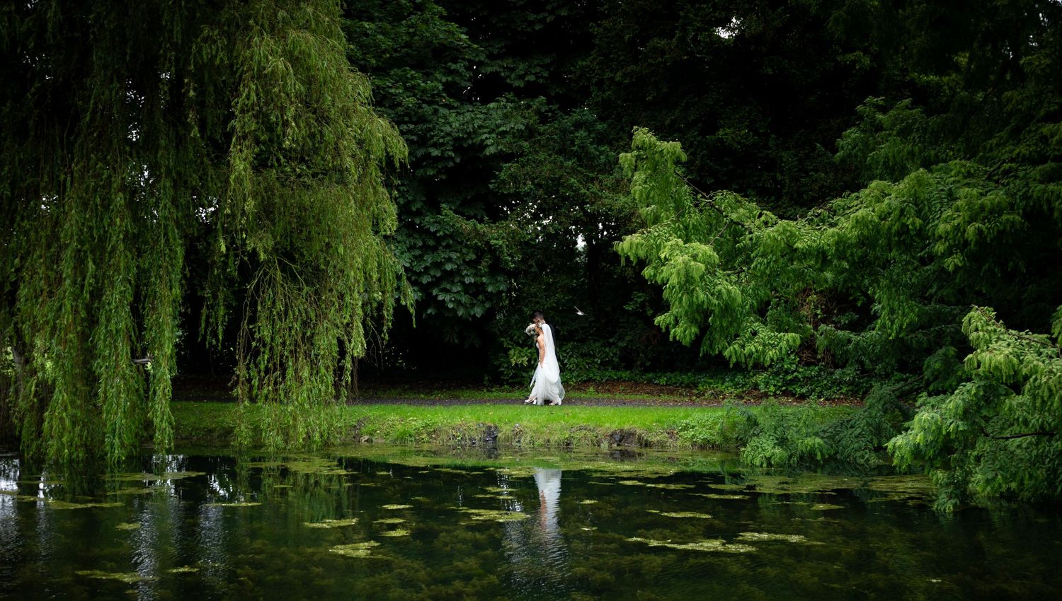Ethereal wedding scene with white dress reflecting in pond surrounded by weeping willows.