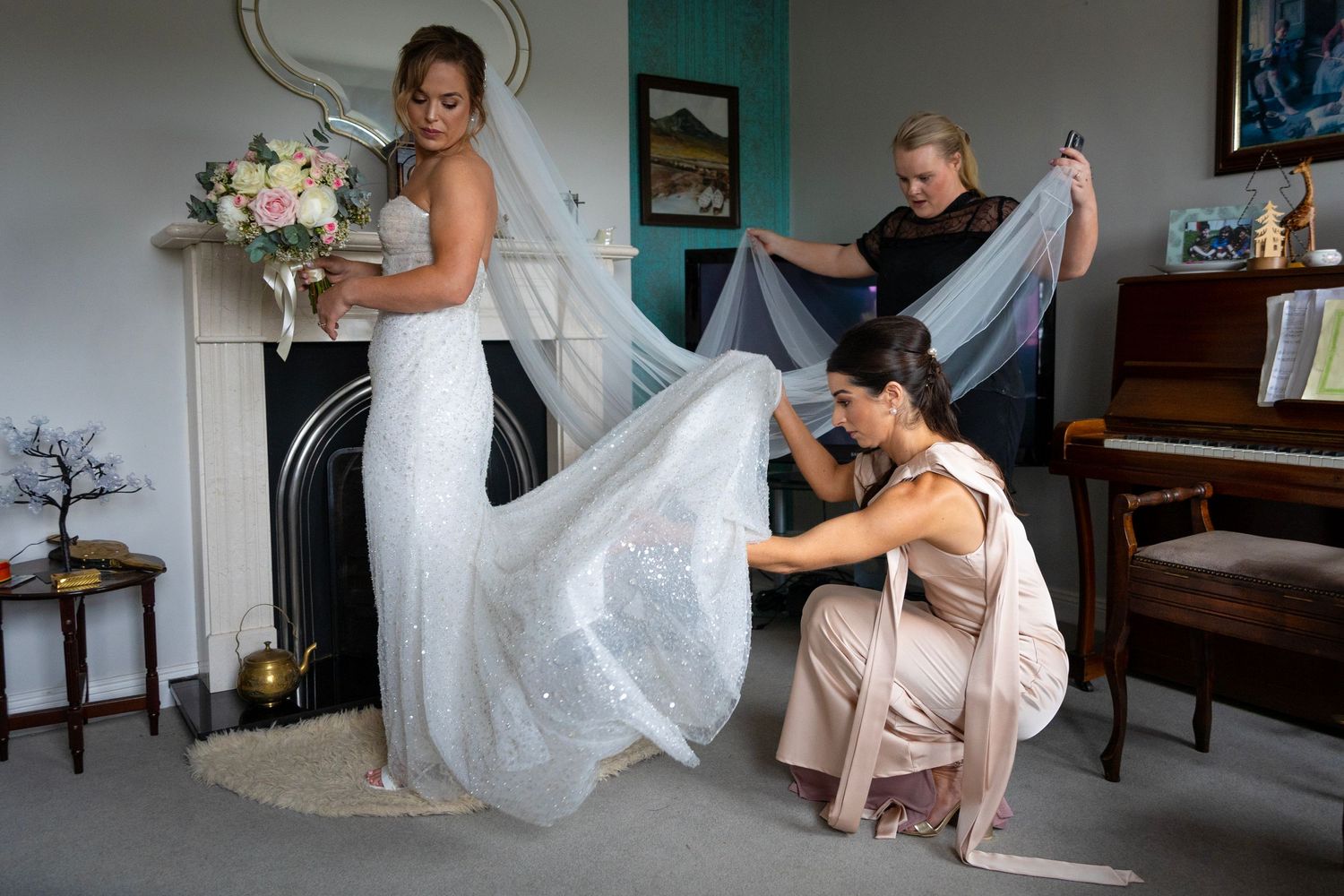Wedding dress preparation scene in elegant room with vintage fireplace and floral arrangements.