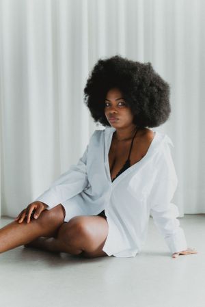 Model with natural afro hair poses in oversized white shirt against light curtain backdrop.
