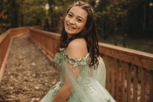 Woman in mint green formal dress poses joyfully on wooden bridge surrounded by autumn foliage.