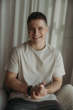 Person in white t-shirt sitting casually and smiling warmly against light curtain background.
