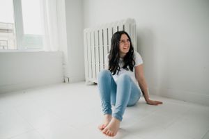 A casual indoor portrait showing someone sitting on a white floor against a radiator wearing jeans and a white t-shirt.