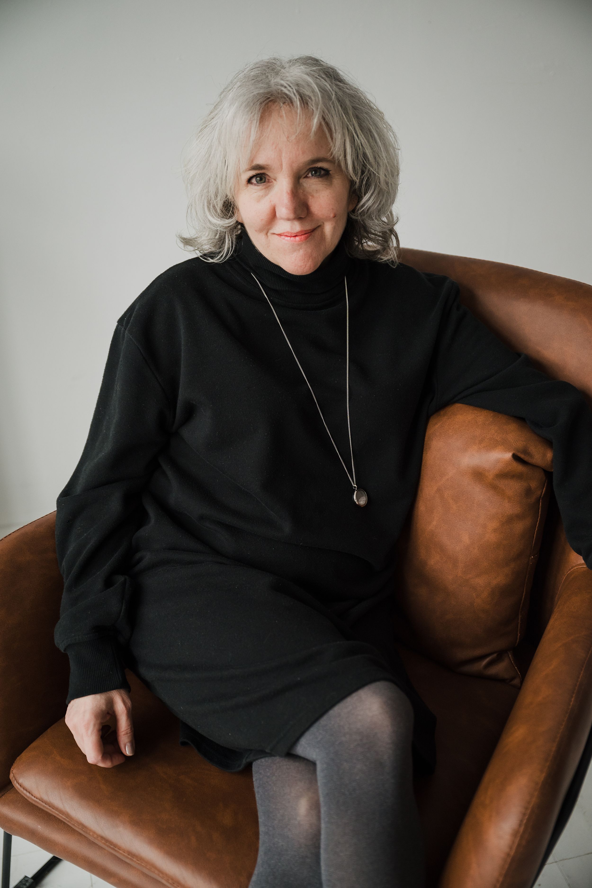 A portrait of someone in a black sweater and necklace sitting on a brown leather chair.