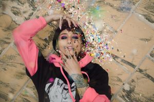 Person lying on ground surrounded by colorful confetti wearing pink and black clothing.