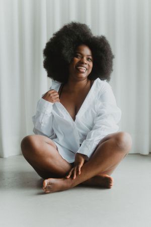 Woman with natural afro hair wearing white shirt sits cross-legged on floor, radiating joy.