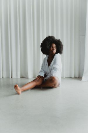 Woman with voluminous afro in white button-down shirt poses on floor against white curtains.