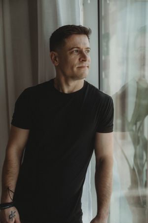 Person in black t-shirt gazes thoughtfully through window with soft natural lighting.