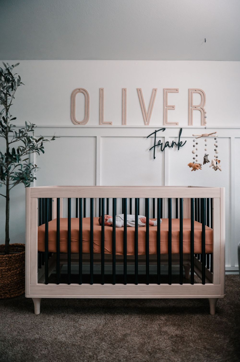Modern white crib with the name 'OLIVER' displayed above in pink letters against a neutral wall.