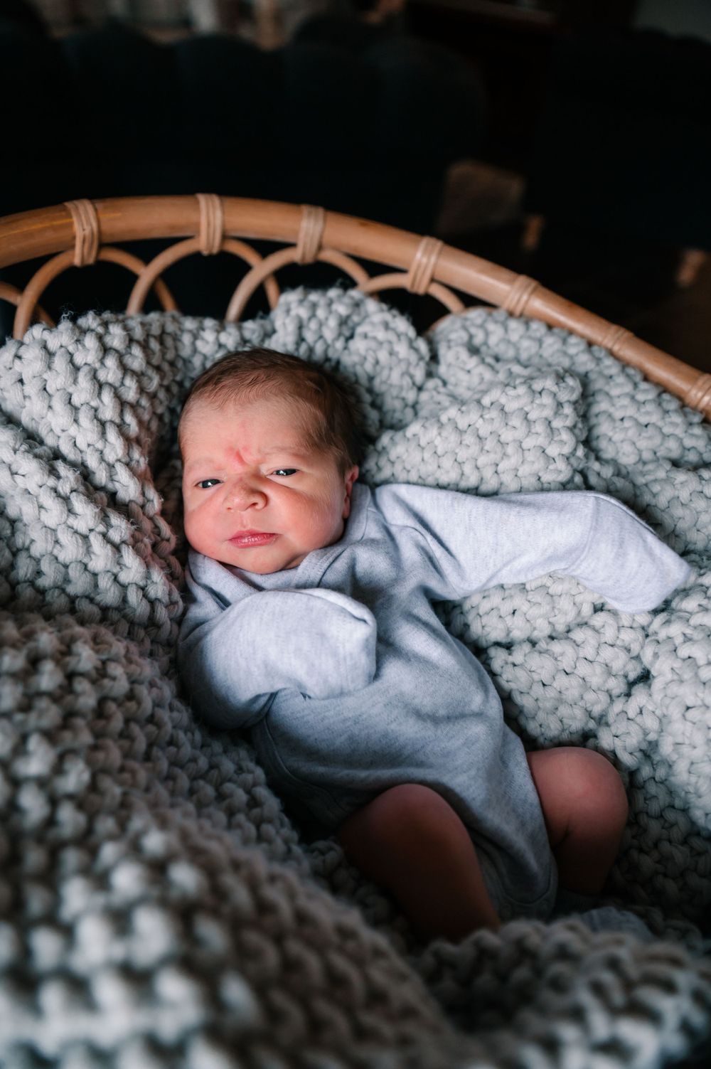 Baby wearing light gray outfit rests on chunky knit blanket in wicker bassinet.