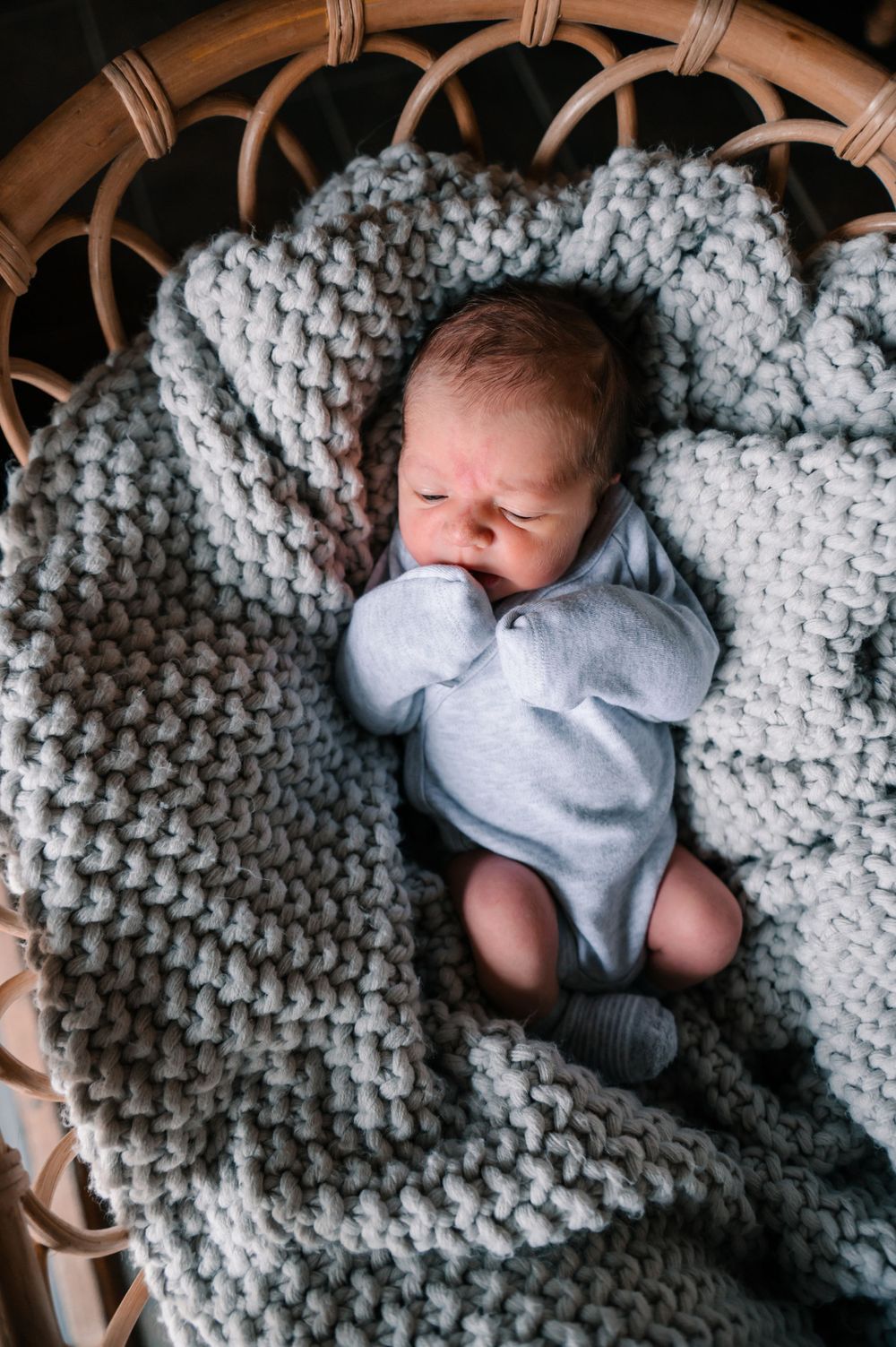 Newborn in gray onesie sleeping peacefully on thick knitted blanket in woven basket.
