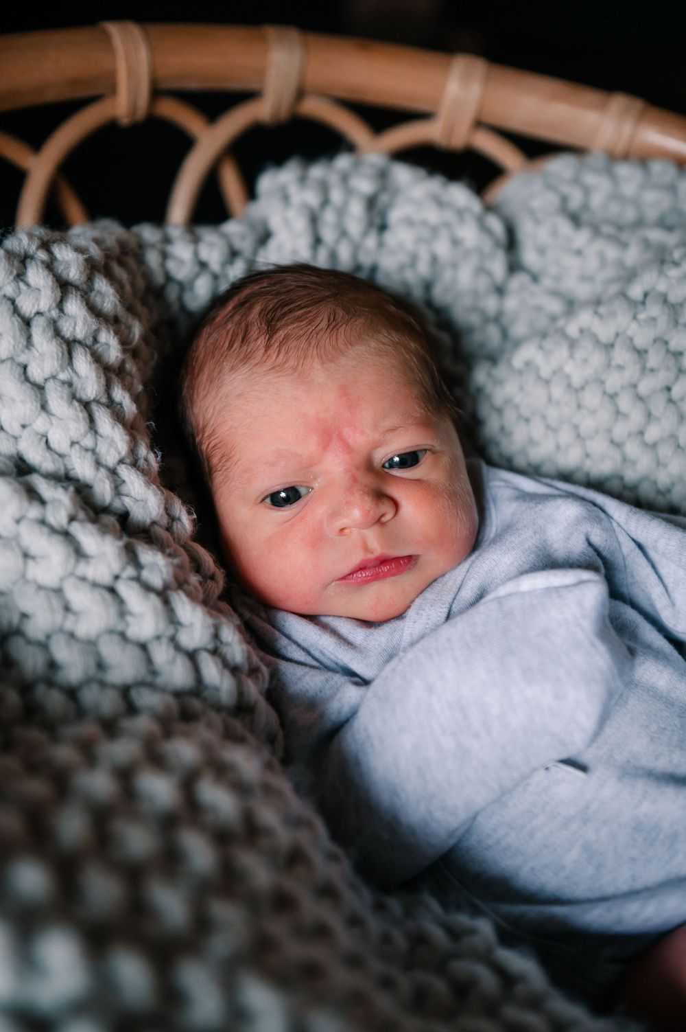 Baby lying on cozy gray knit blanket in rattan bassinet with soft lighting.