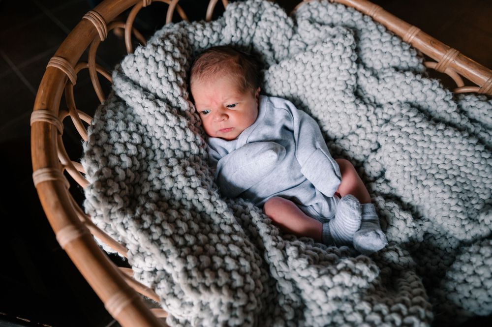 Infant resting in wicker bassinet covered with chunky knit gray blanket.