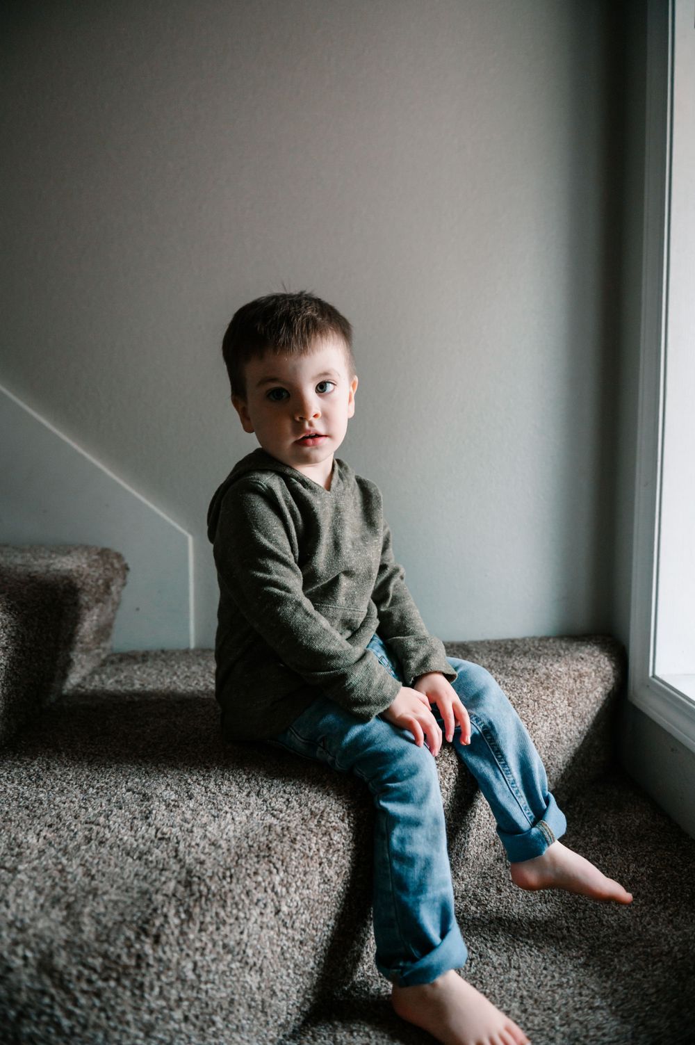 Child in gray sweater and jeans sitting by window on carpeted floor.