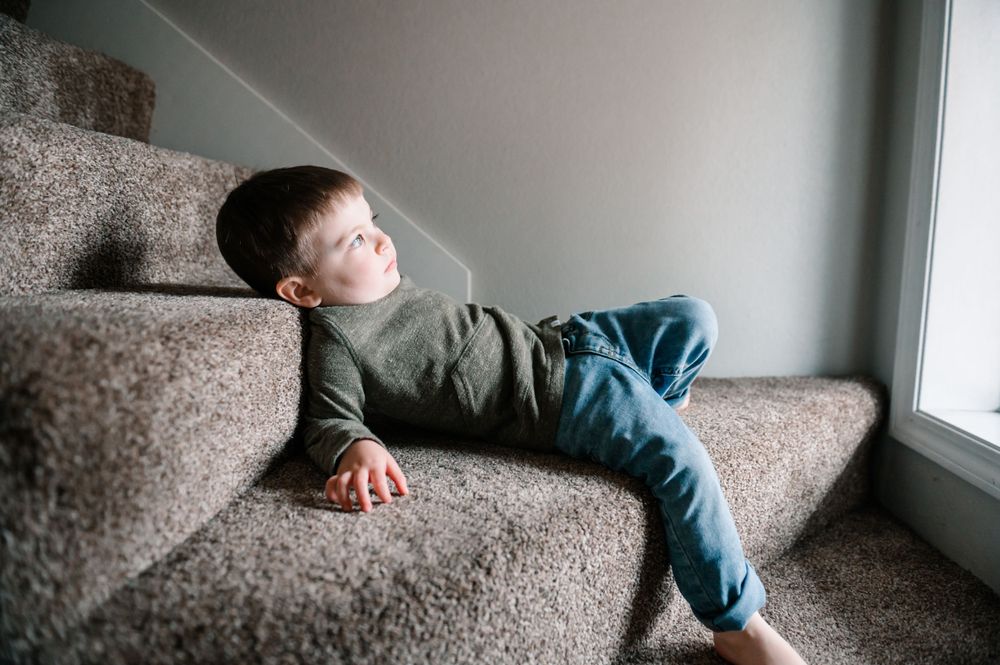 Young child relaxing on gray carpet near window in casual attire.