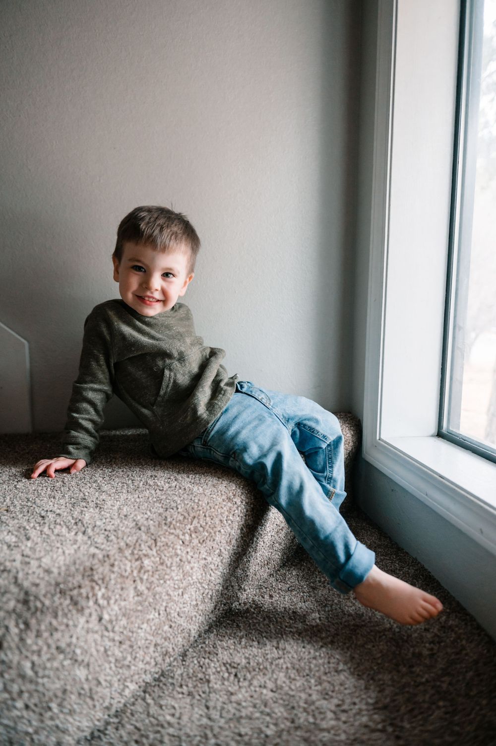 Child playing and smiling near window on carpeted floor wearing casual clothes.
