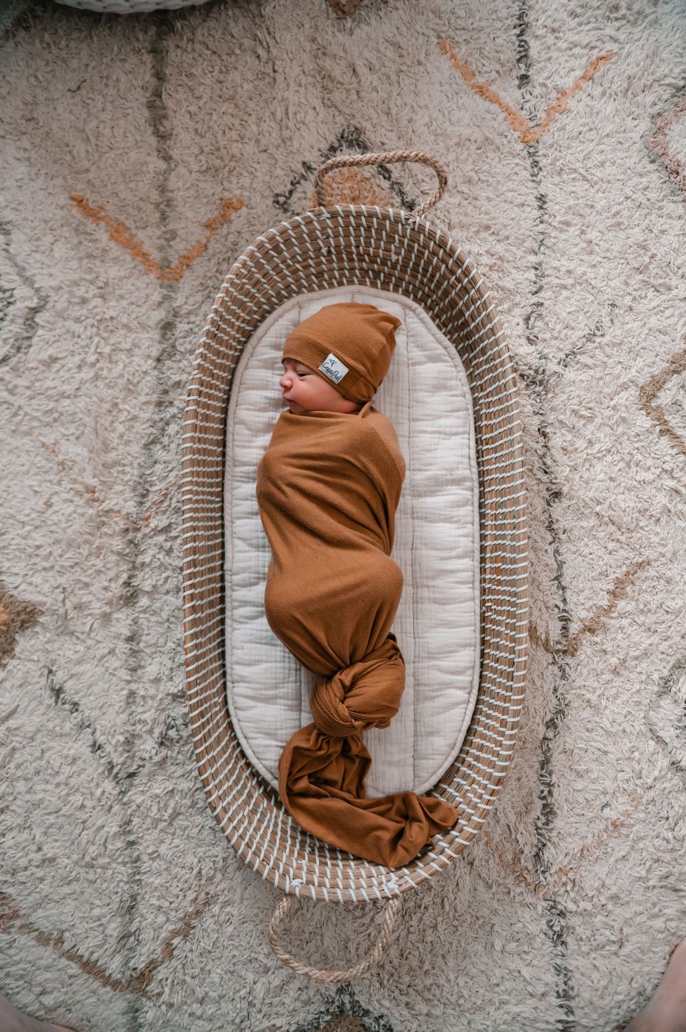 Brown swaddled newborn lying in oval-shaped wicker bassinet on textured rug.