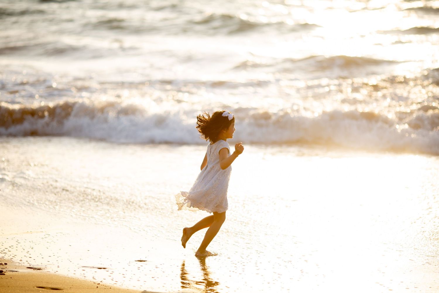 Child in white dress running joyfully on beach at golden sunset.