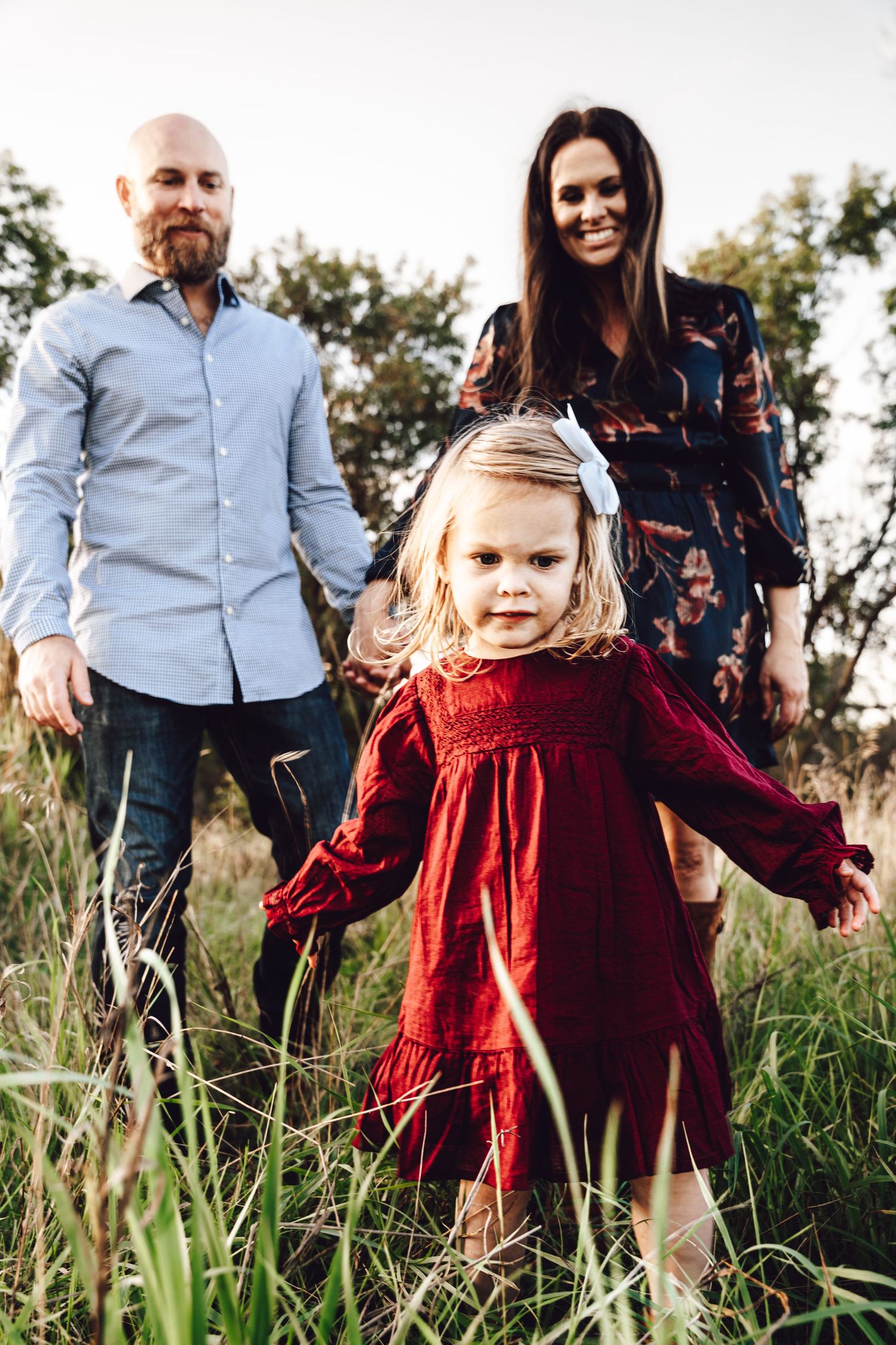 Family portrait in tall grass field during golden hour with child in red dress.