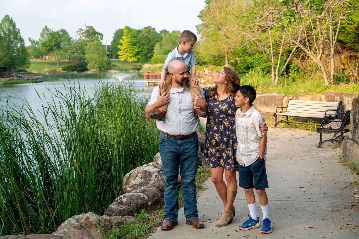 Happy family of four walking along a scenic park path near a pond surrounded by lush greenery.